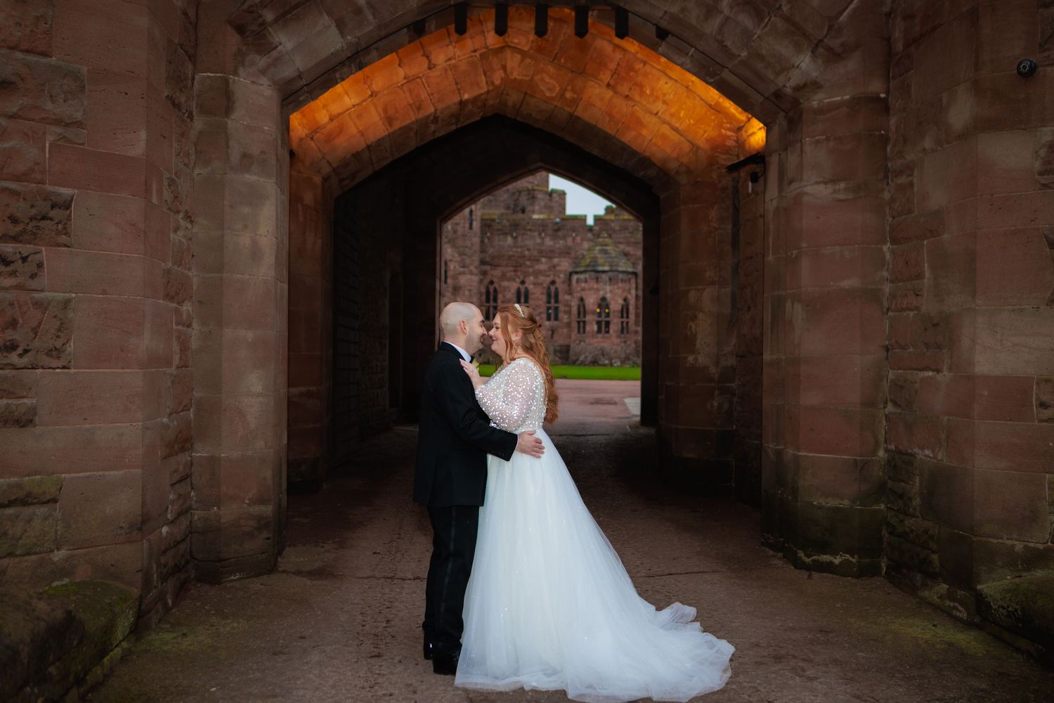 A couple embraces in a stone archway passage at dusk with a historic building visible in the background.
