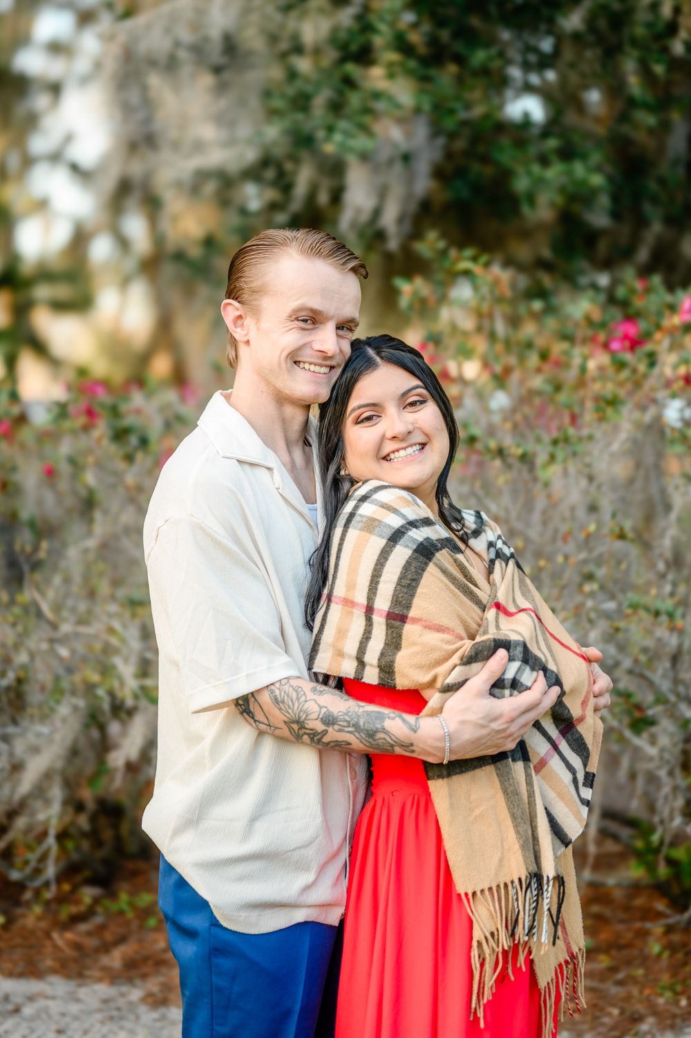 A couple embraces outdoors wearing a red dress and plaid scarf against a natural backdrop with Spanish moss.