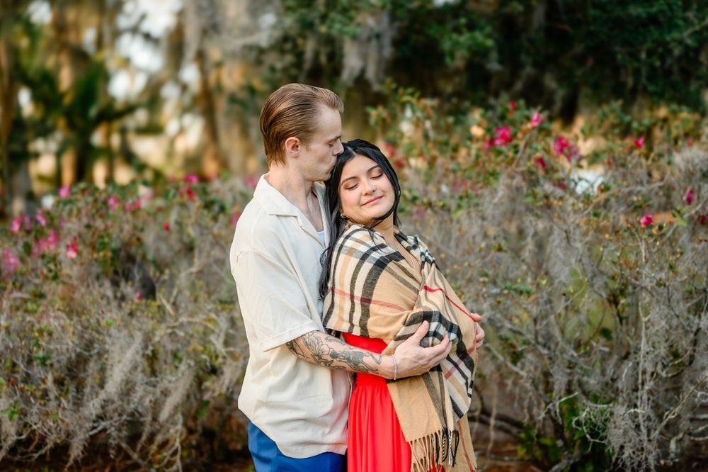 A couple shares a romantic moment in a garden setting with flowering plants and trees in the background.