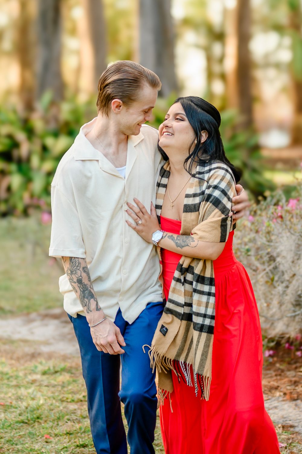 A romantic couple shares a tender moment in a forest setting, with her wearing a red dress and plaid scarf.