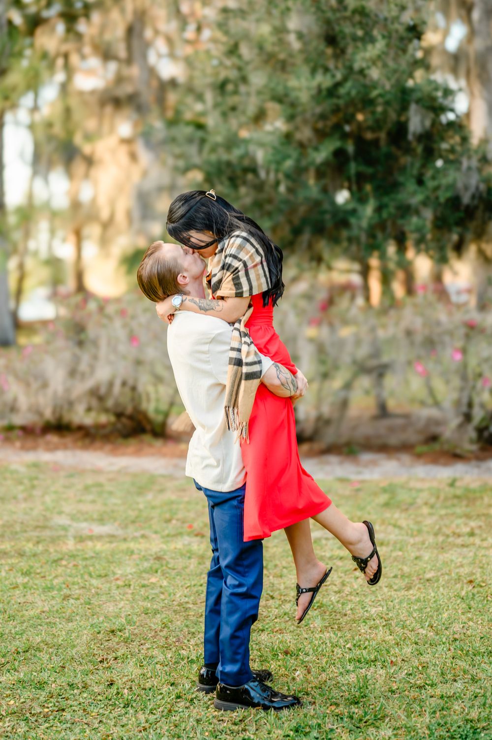 A romantic couple shares a playful moment as he lifts her up in a garden setting with Spanish moss trees.