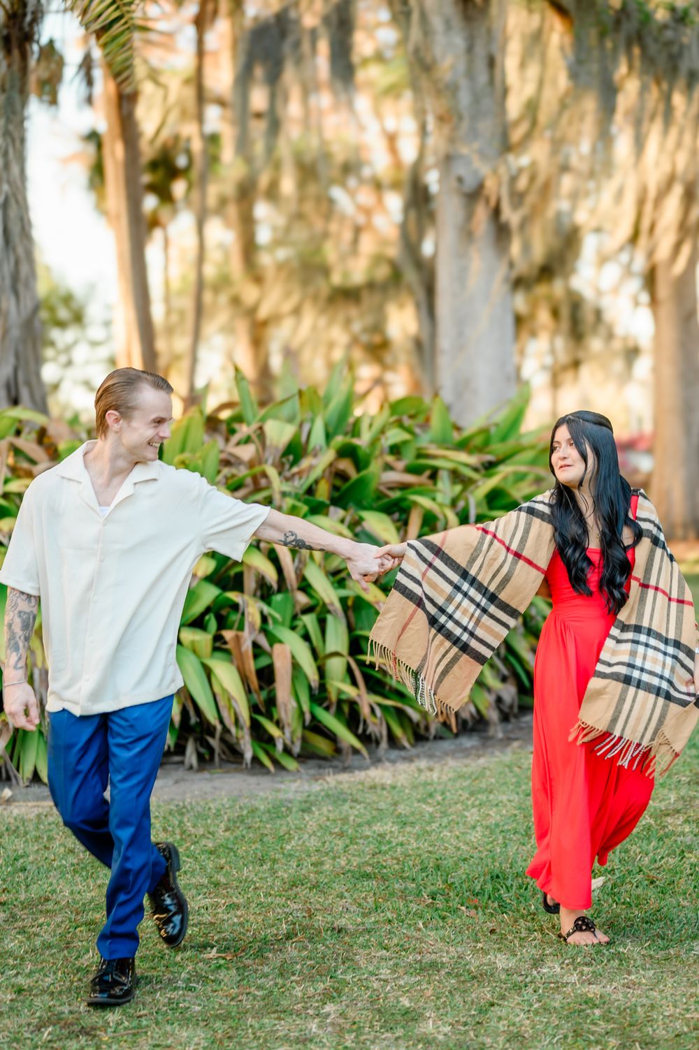 A couple dances playfully on grass with Spanish moss trees in background, she wearing a red dress and plaid shawl.