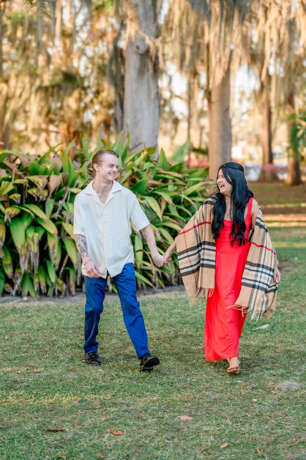 A couple dances playfully on grass with trees and foliage creating a natural outdoor backdrop.