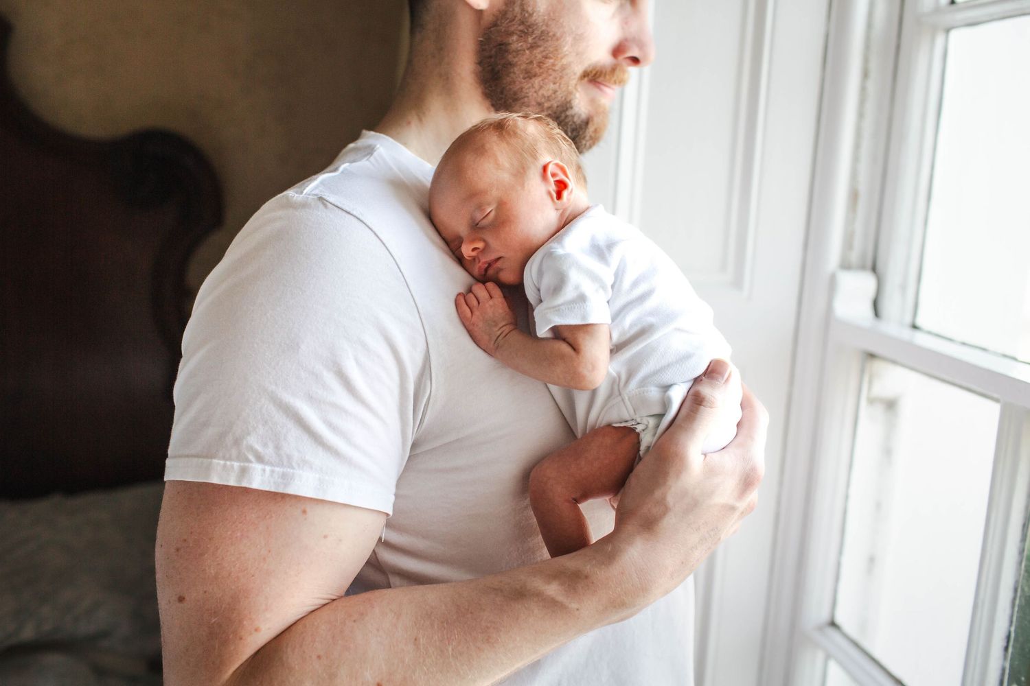 A tender moment between parent and newborn baby in white clothing by a bright window.