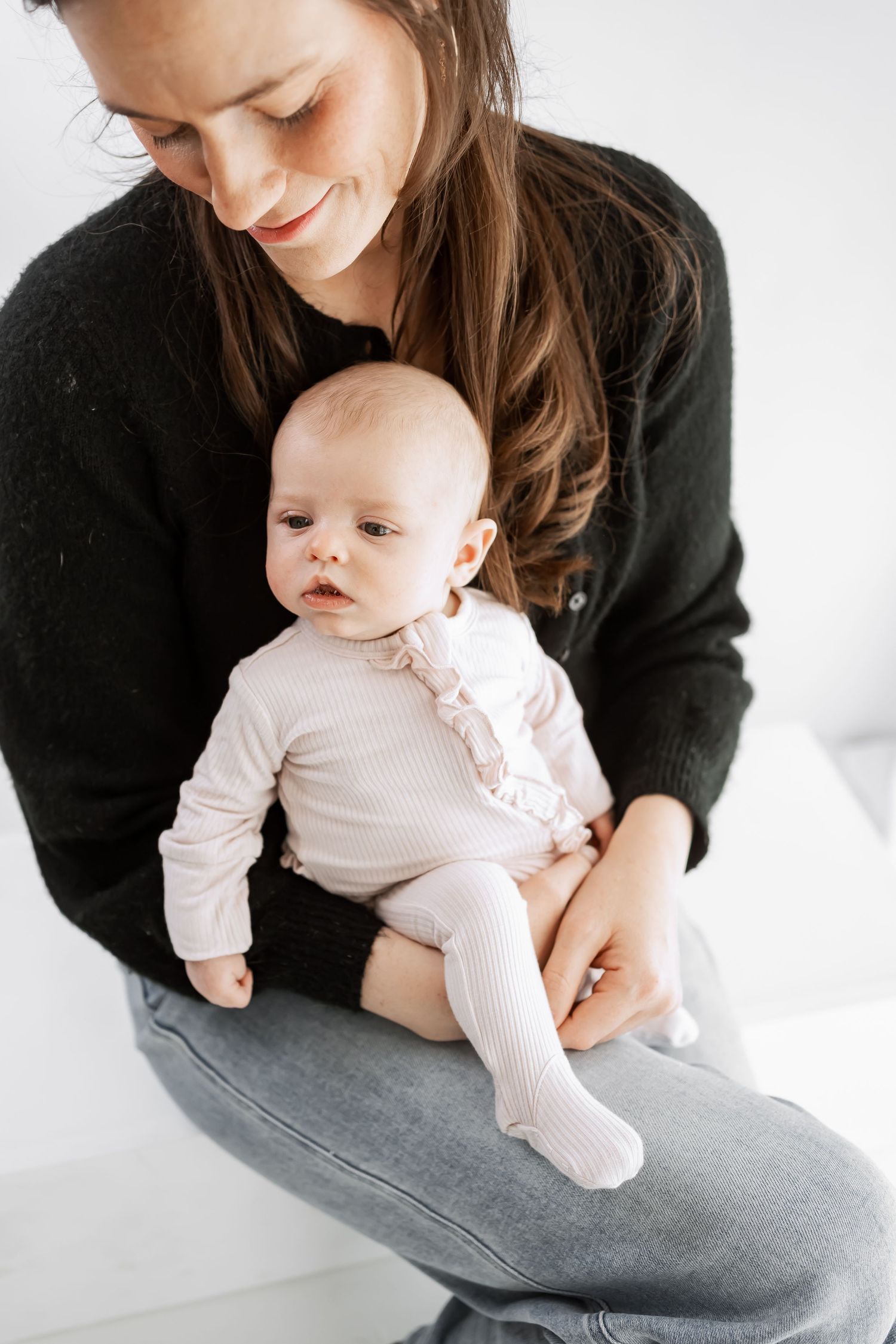A tender moment between parent and baby wearing white, sitting together in casual attire against a light background.