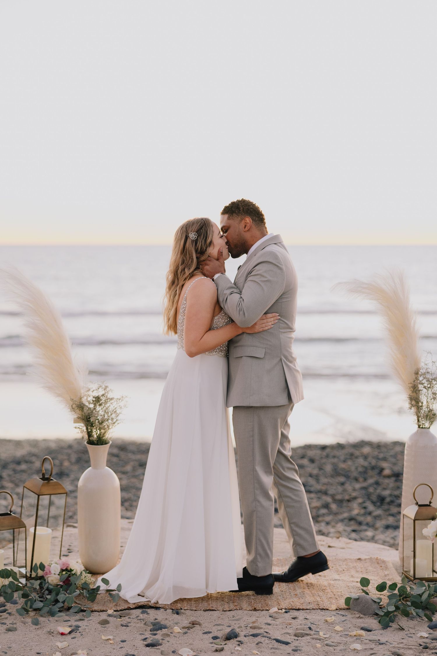 Smiling newlyweds hold hands strolling along a serene beach at golden hour with bouquet.