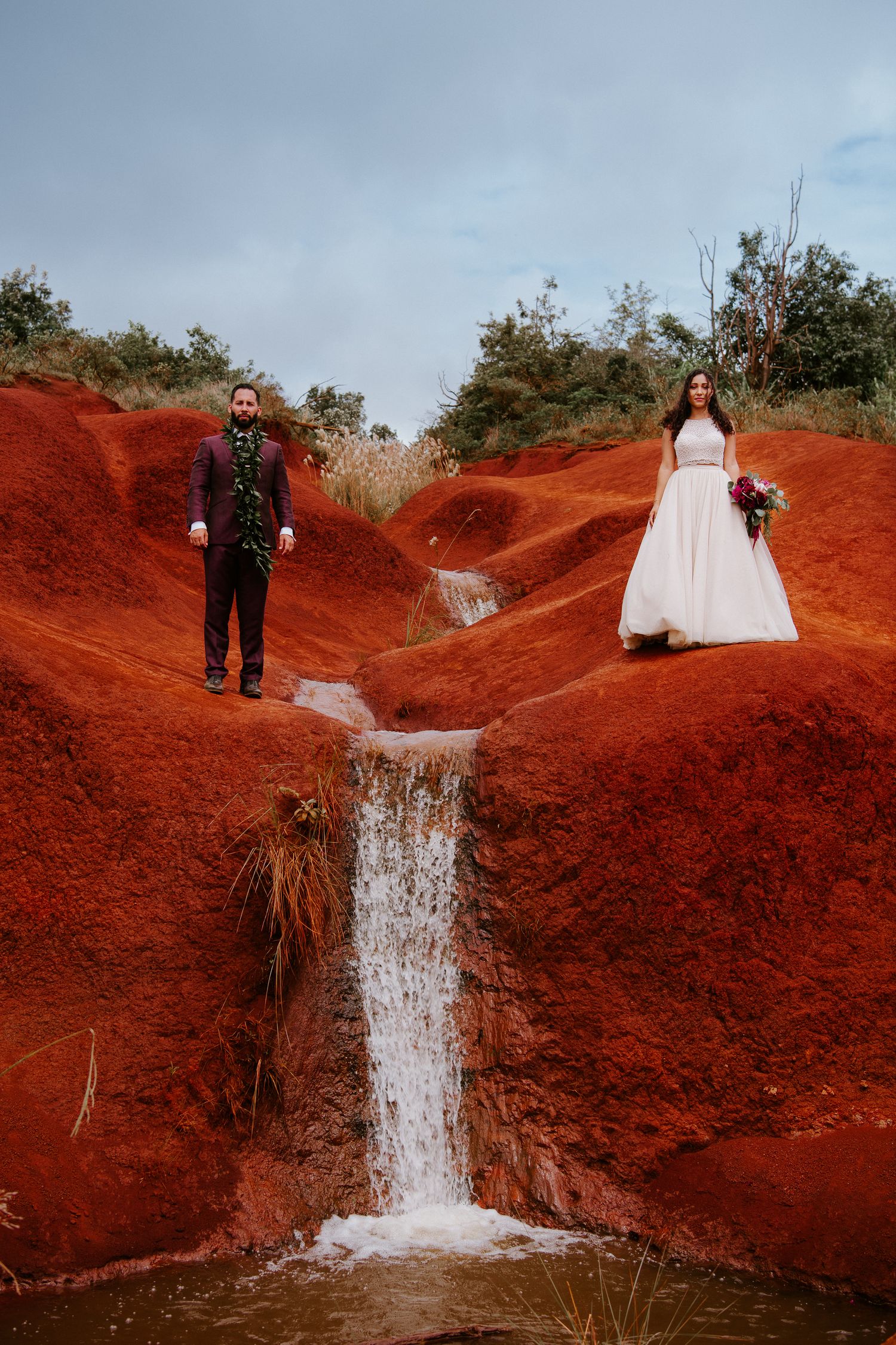 Dramatic wedding photo of bride and groom standing on vivid red volcanic terrain beside a small waterfall.