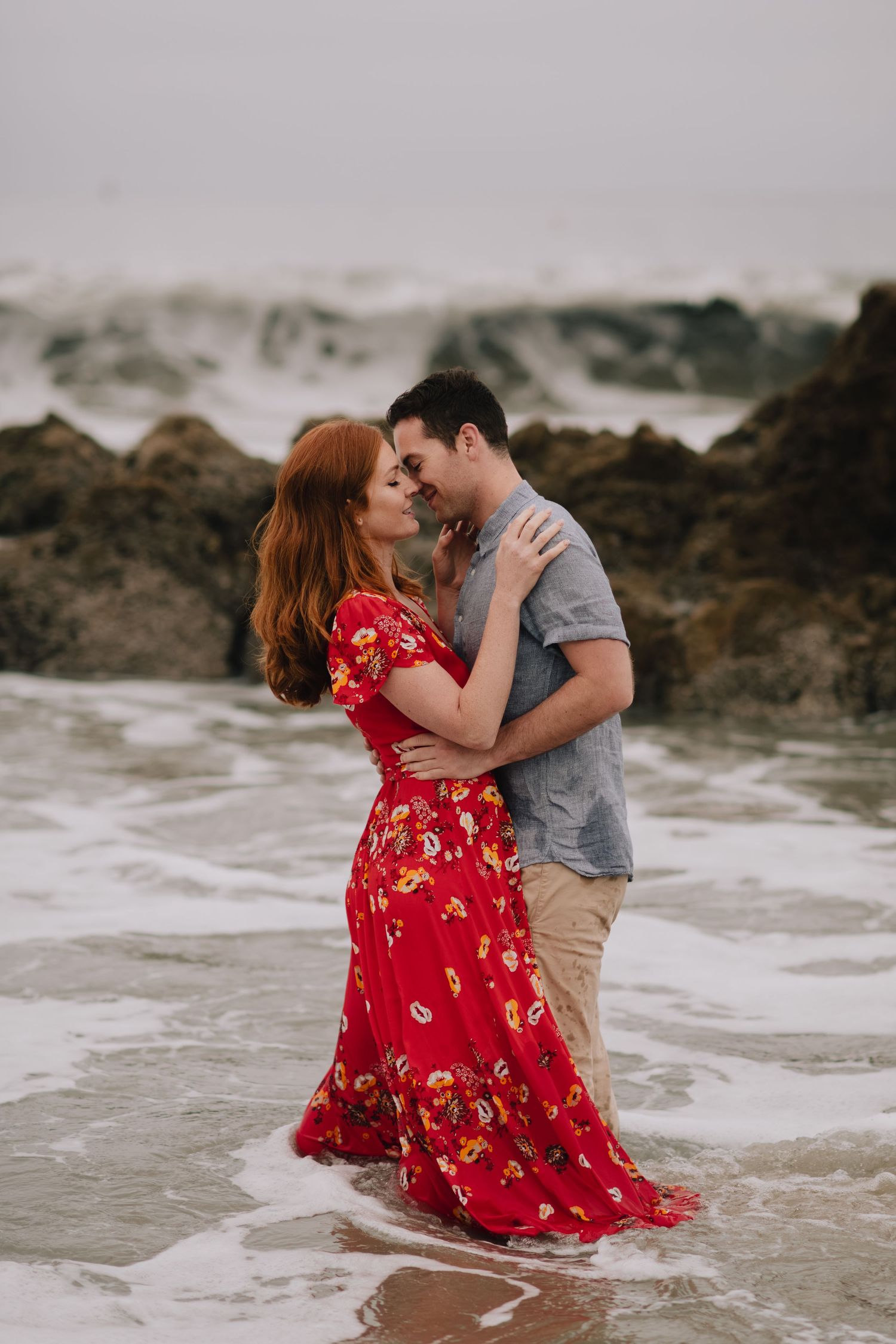 Couple romantically embracing in ocean surf, woman in flowing red floral maxi dress, rocky coastline.