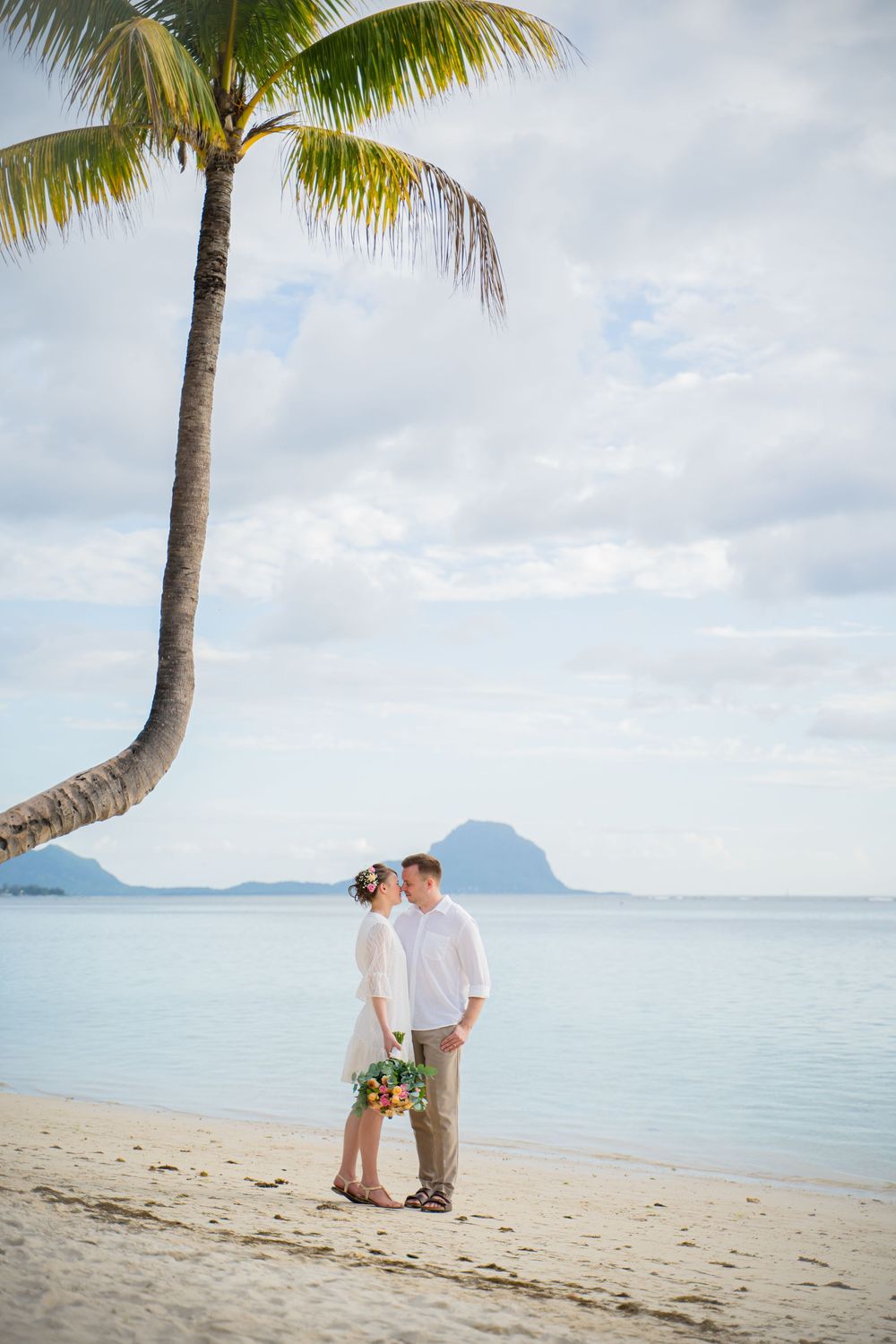A couple stands on a tropical beach with a leaning palm tree and distant mountains over calm turquoise waters. Shot by Luminair Studio