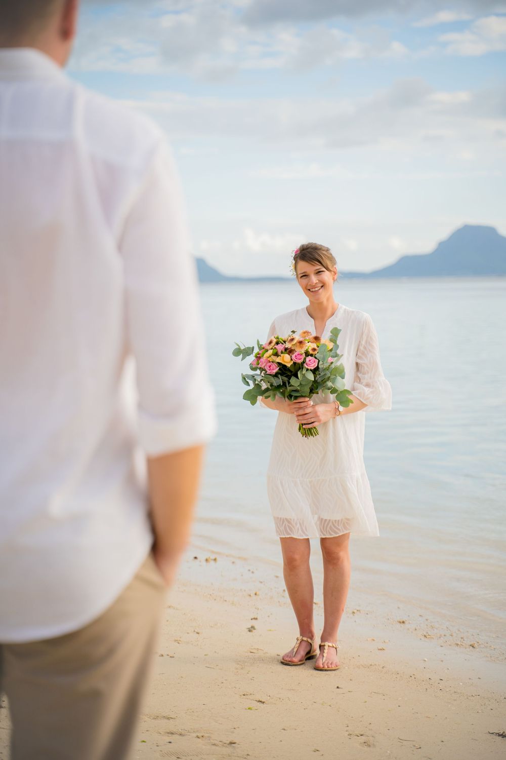 A couple shares an intimate moment on a pristine beach with turquoise waters and mountains in the background. Shot by Luminair Studio