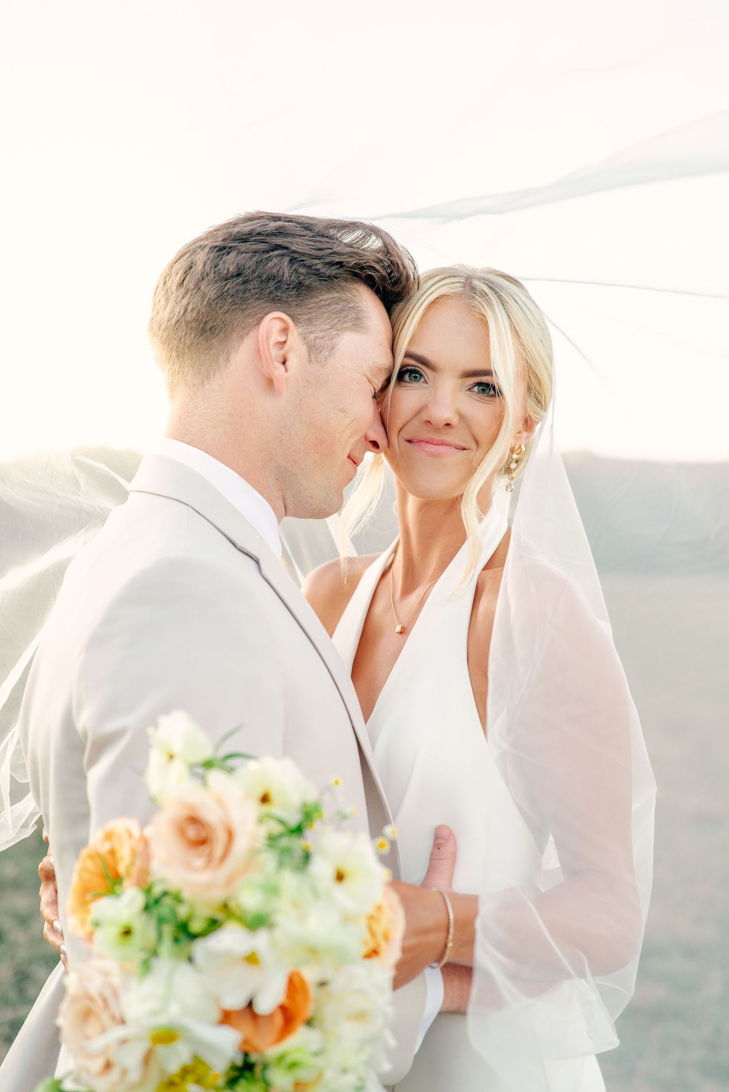 A romantic outdoor wedding photo with a bride in white holding a peach and white bouquet during a sun-filled moment.
