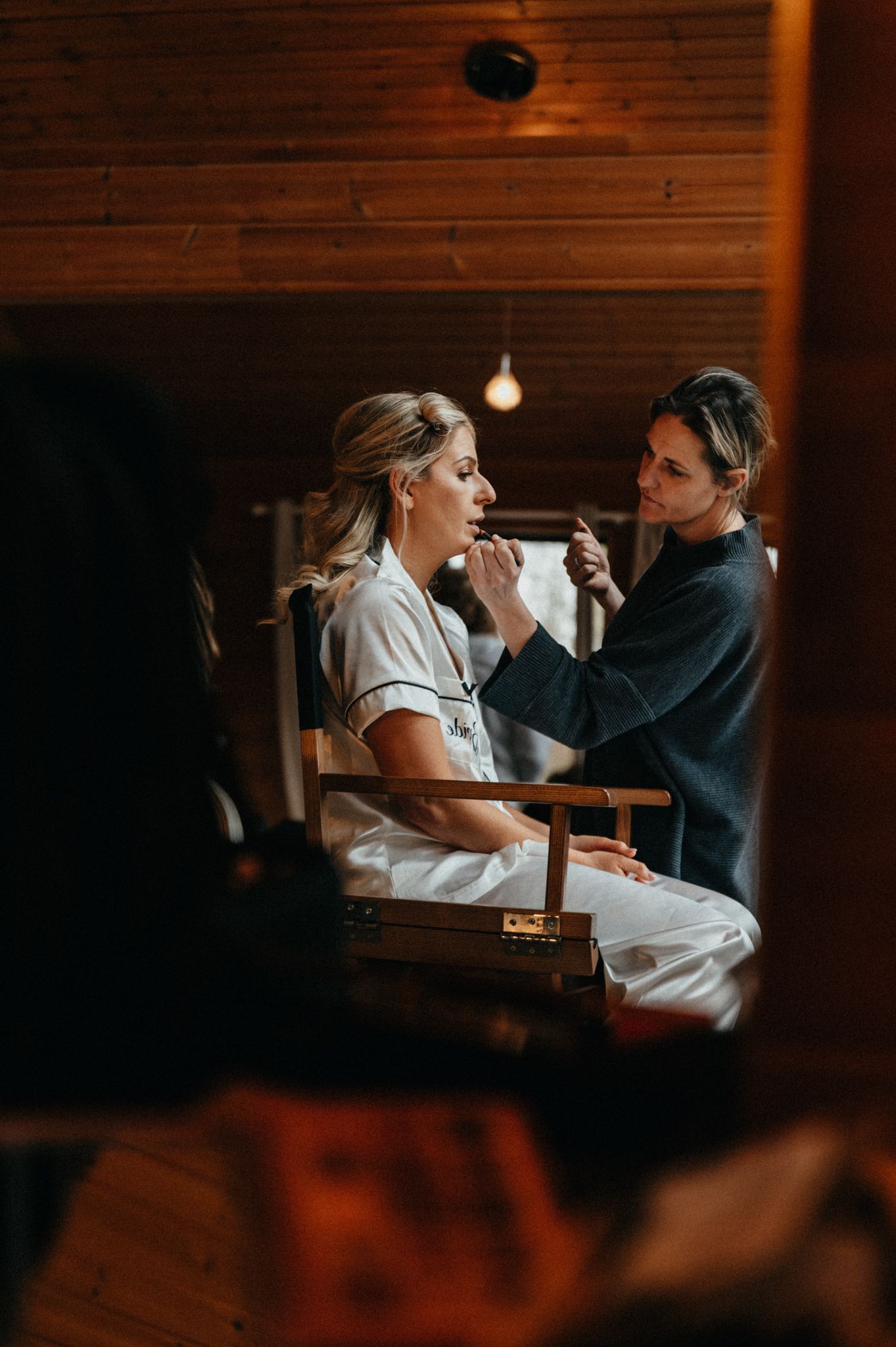 A makeup artist applies cosmetics in a warmly lit wooden room during wedding preparation.