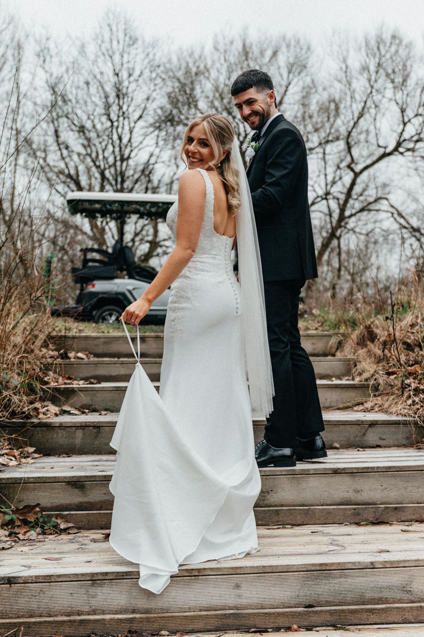 A couple in wedding attire walks up wooden steps together on their special day in a rustic outdoor setting.