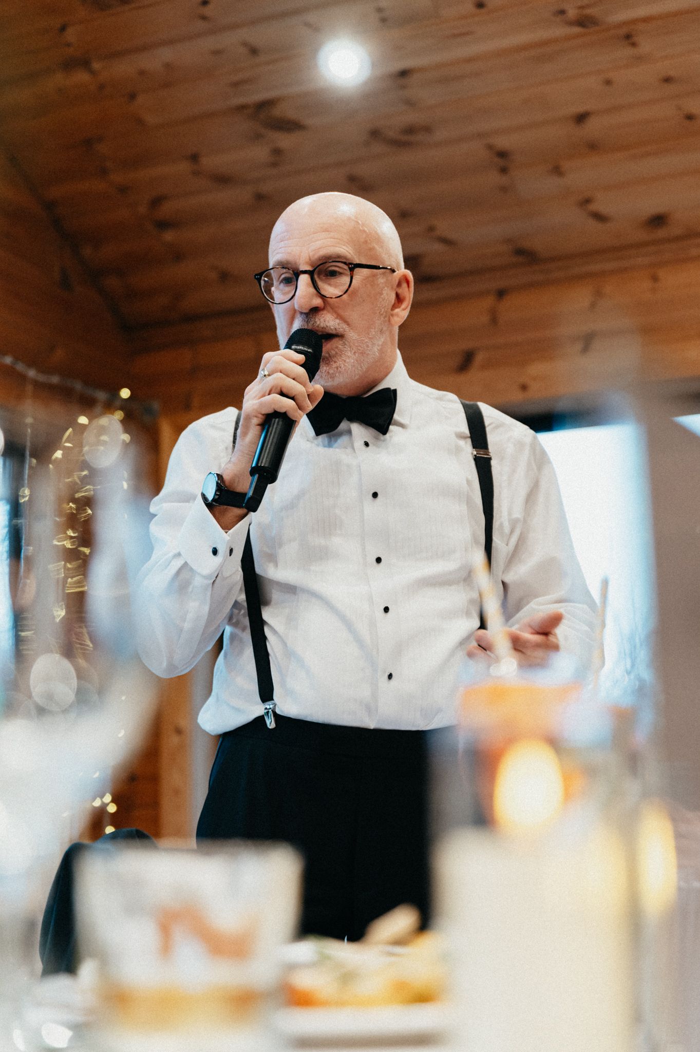 Someone in a white dress shirt, bow tie and suspenders speaks into a microphone during an elegant indoor event.
