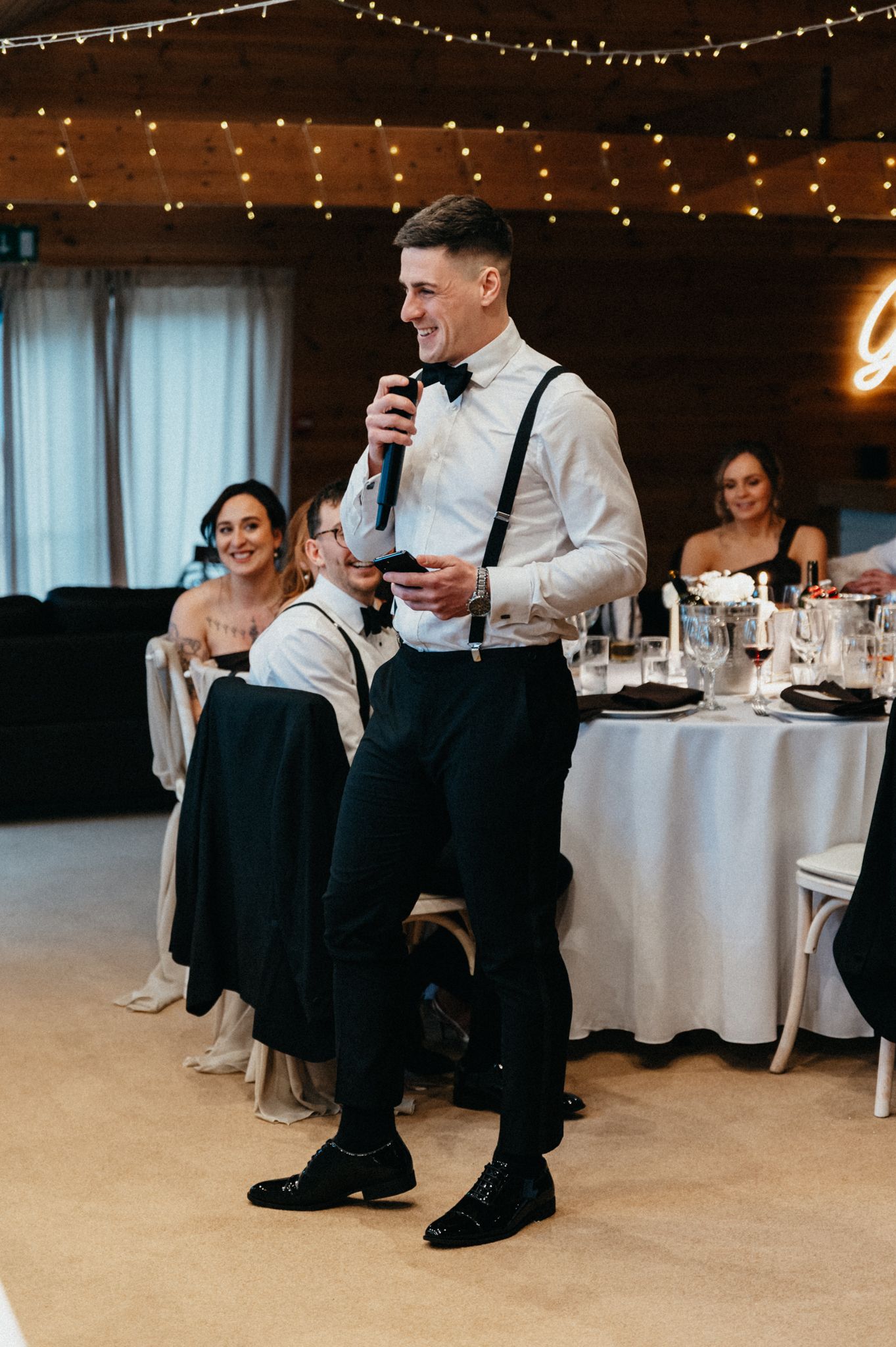 Wedding reception speech being given by someone in suspenders and white shirt at elegant table setting.