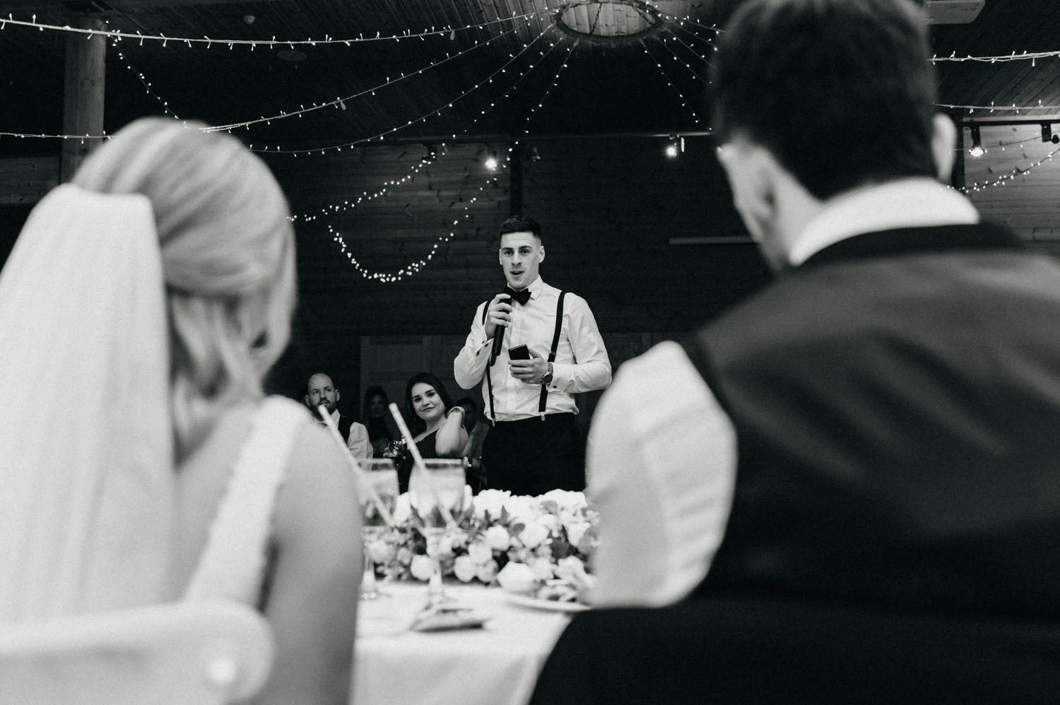 A speaker delivers a wedding toast at the reception while guests listen from decorated tables.