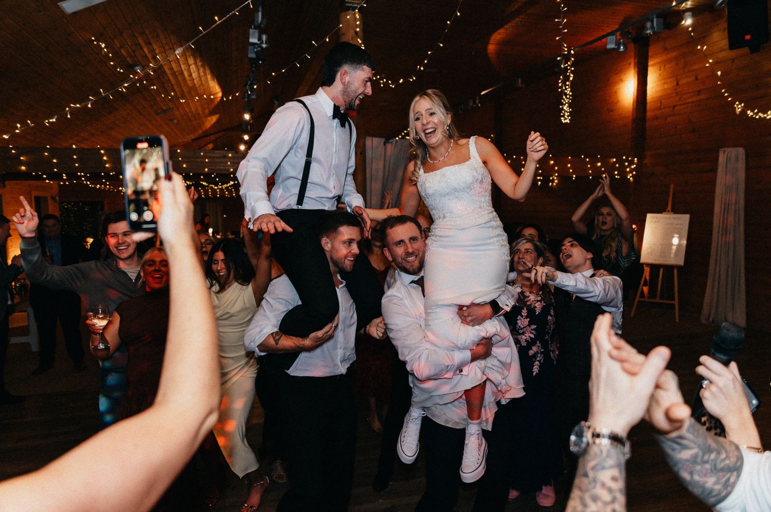 Wedding guests celebrate on the dance floor under string lights while taking photos with their phones.