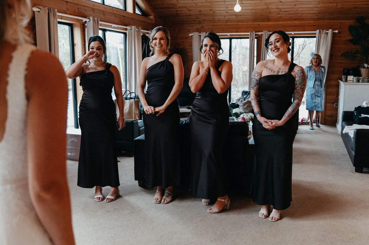 Wedding party members in black formal attire gather emotionally in a rustic wooden venue.