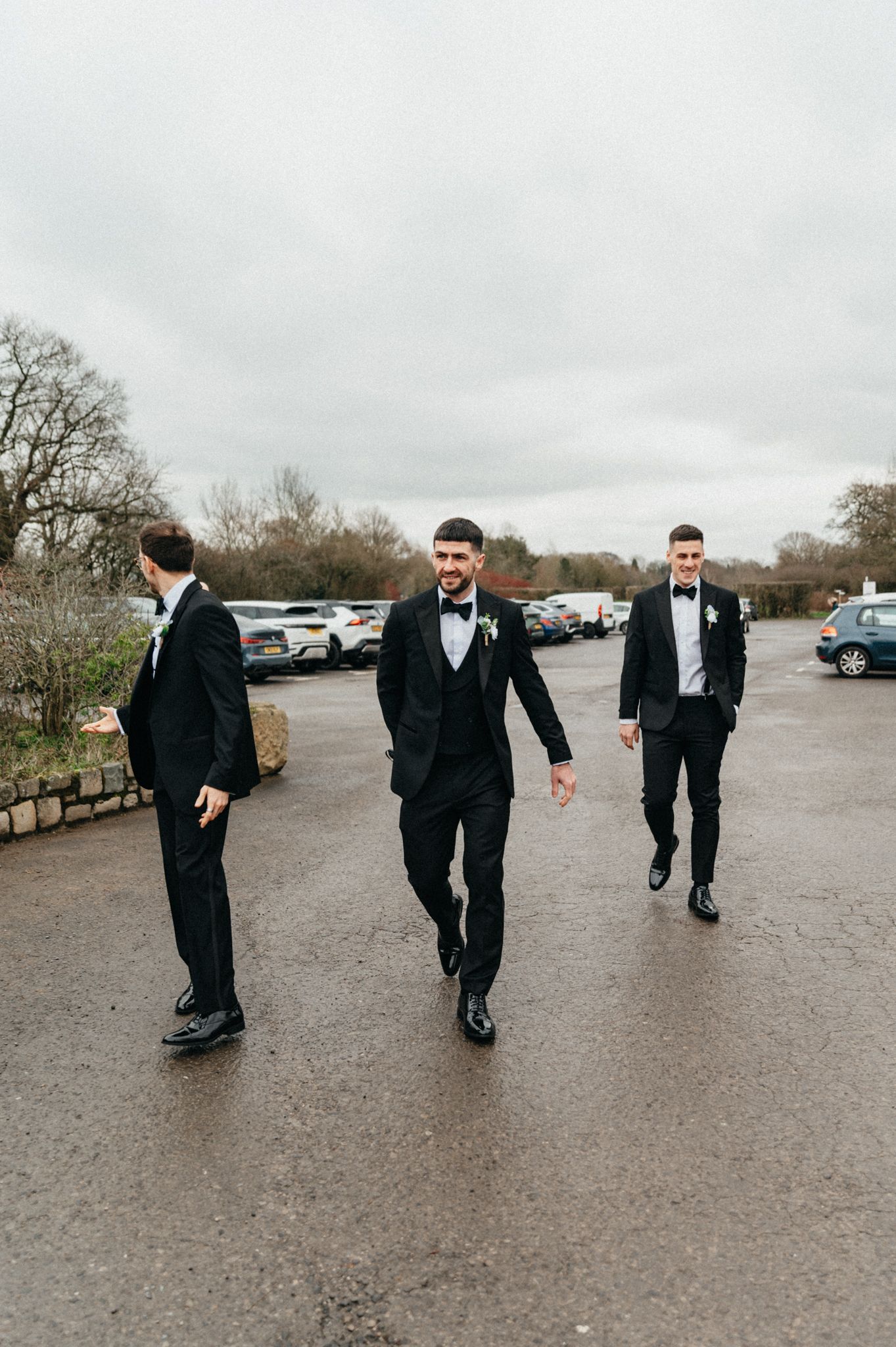 Groomsmen in black tuxedos walking playfully across a wet parking lot on a cloudy day.