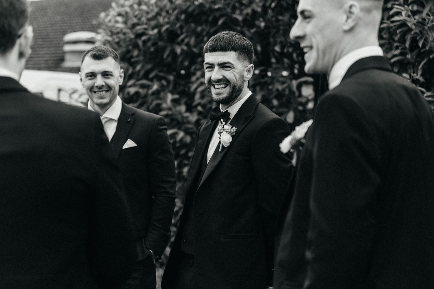 Wedding party members in formal black suits smiling and laughing together outdoors.