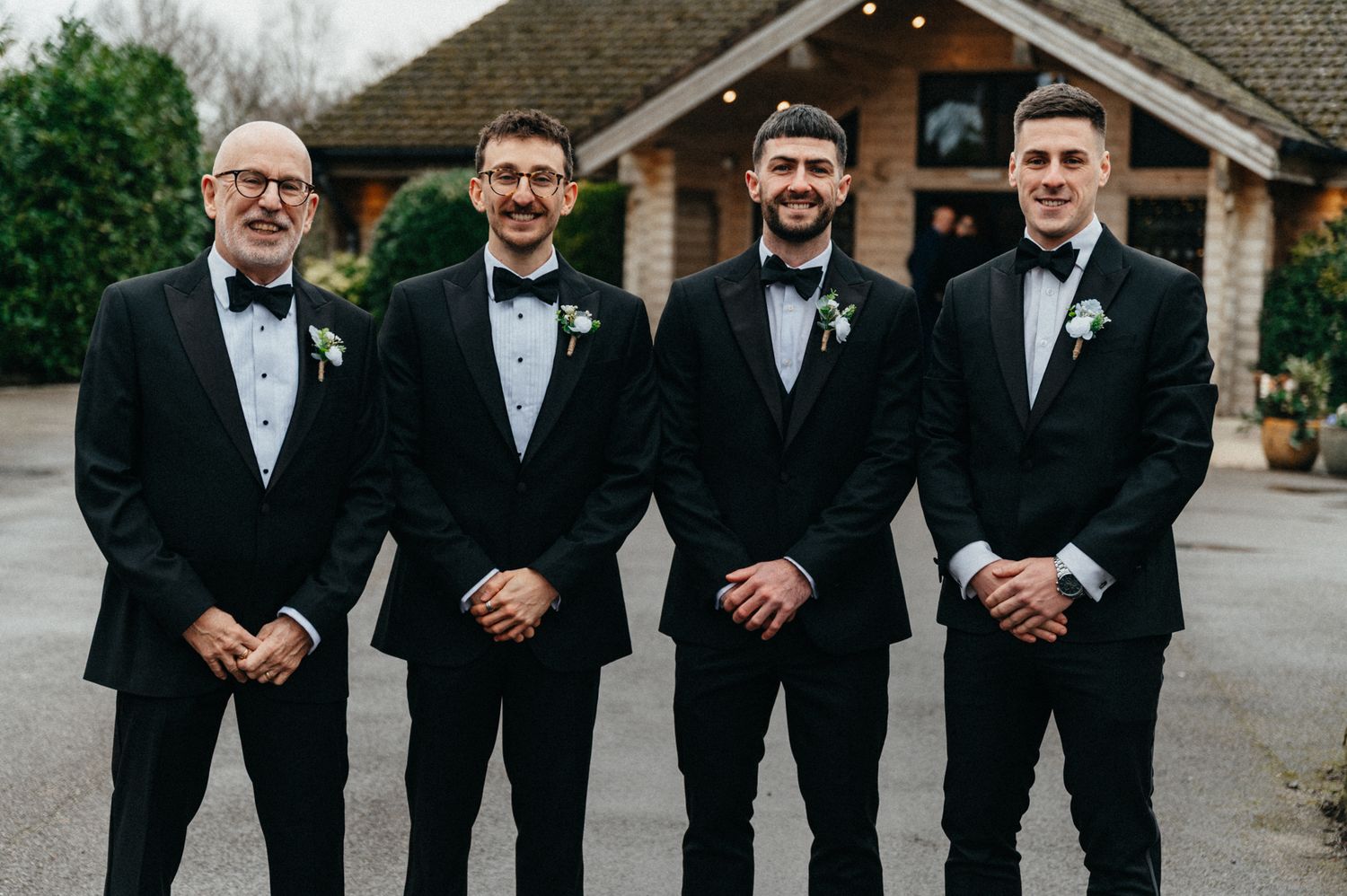 Groomsmen in black tuxedos and bow ties lined up for formal wedding photos outside.