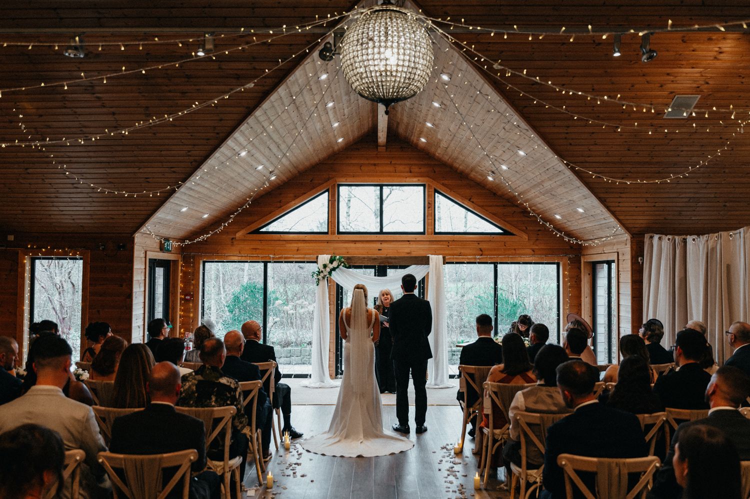 Indoor wedding ceremony in a rustic wooden venue with vaulted ceiling and twinkling string lights overhead.