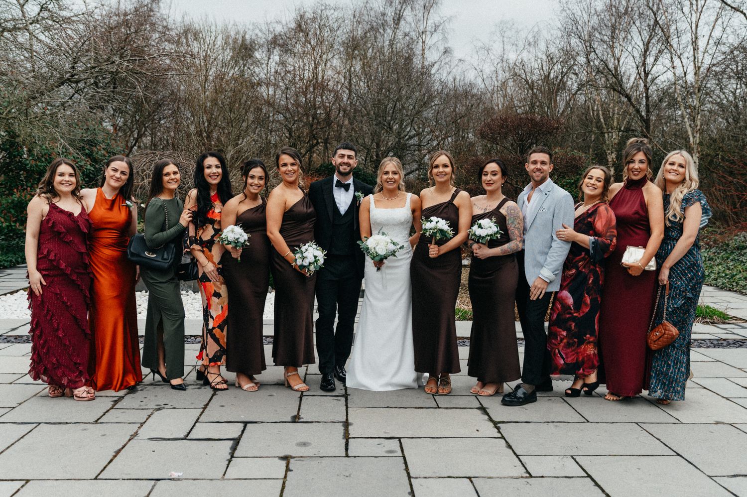 Wedding party group photo showing bridesmaids in burgundy and black dresses lined up outdoors.