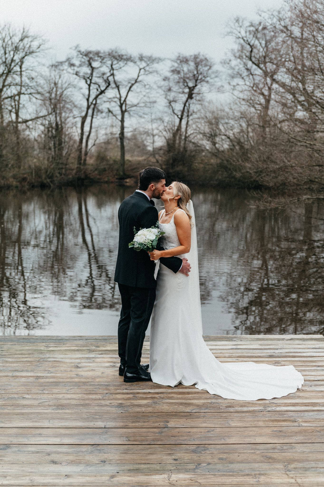 A couple shares a romantic moment on a wooden dock overlooking a serene lake surrounded by bare winter trees.