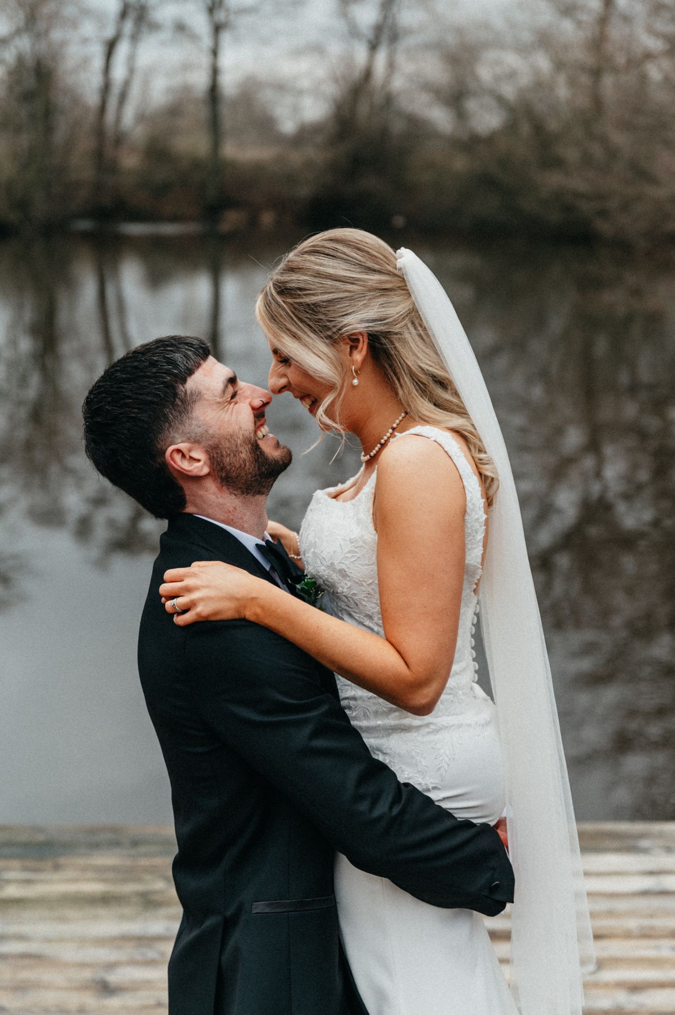 A romantic wedding couple embraces by a serene lake on their special day, with a flowing white veil and formal attire.