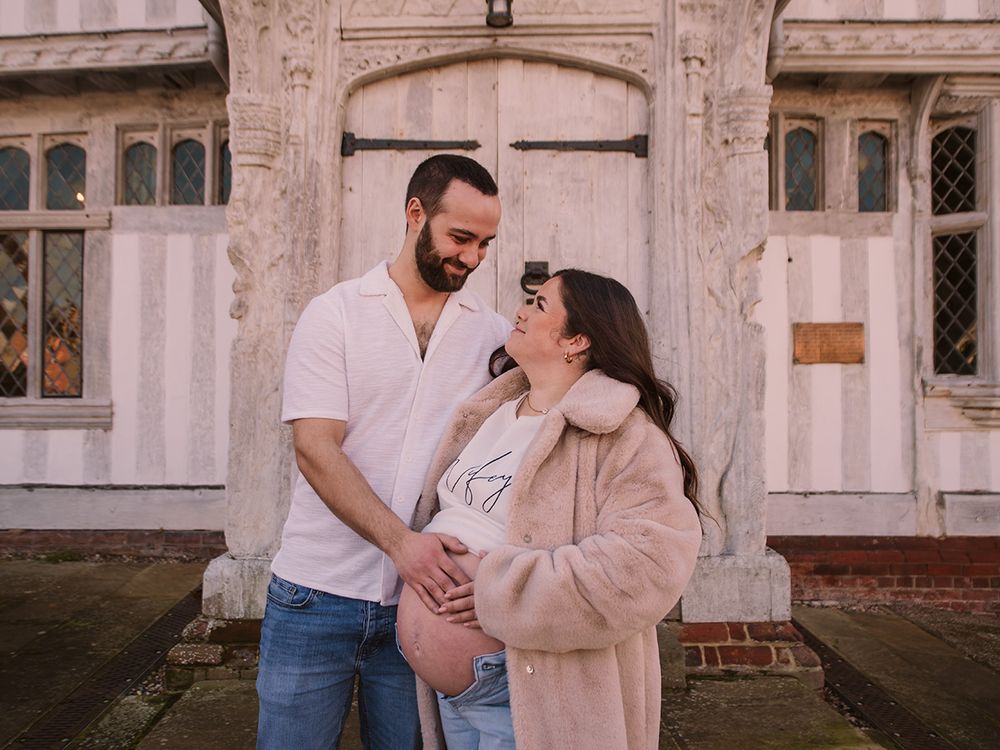 Mum‑to‑be in jeans and jumper cradling her bump in historic Lavenham market square, by all scene images photography. Essex, UK