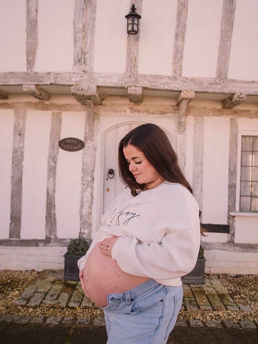 Mum‑to‑be in jeans and jumper cradling her bump in historic Lavenham market square, by all scene images photography. Essex, UK