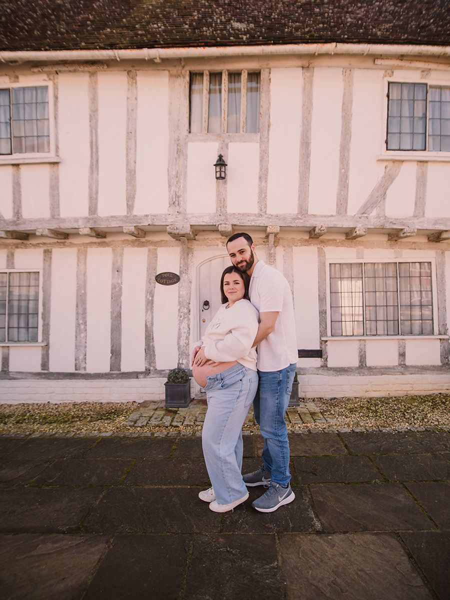 Mum‑to‑be in jeans and jumper cradling her bump in historic Lavenham market square, by all scene images photography. Essex, UK