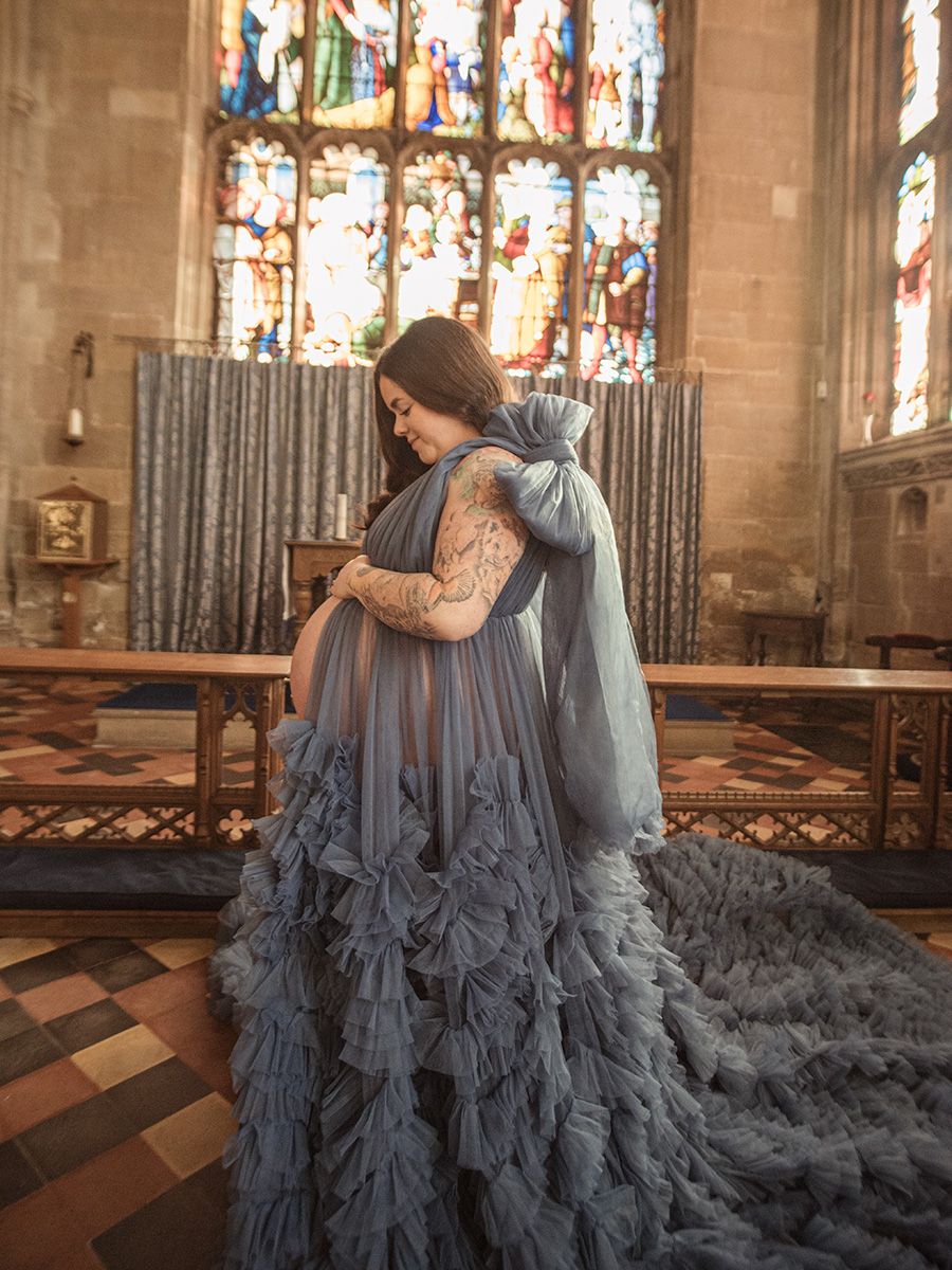 Mum‑to‑be in flowing gown cradling her bump in historic Lavenham St Peter & St Pauls Church, by all scene images photography, Essex, UK