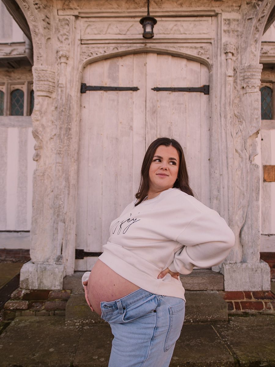 Mum‑to‑be in jeans and jumper cradling her bump in historic Lavenham market square, by all scene images photography. Essex, UK