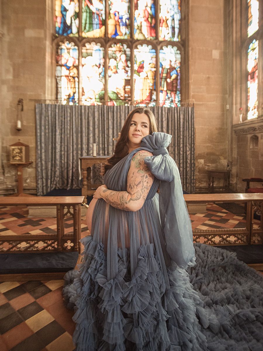 Mum‑to‑be in flowing gown cradling her bump in historic Lavenham St Peter & St Pauls Church, by all scene images photography, Essex, UK