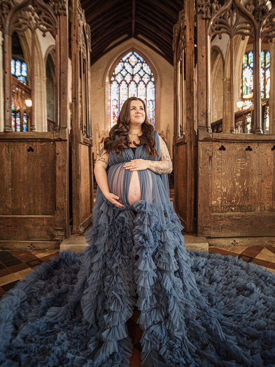 Mum‑to‑be in flowing gown cradling her bump in historic Lavenham St Peter & St Pauls Church, by all scene images photography, Essex, UK