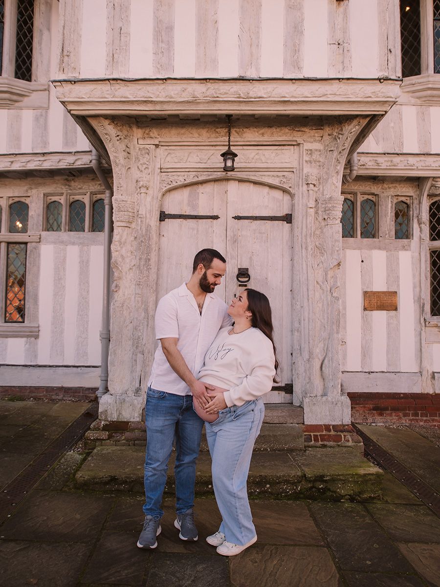 Mum‑to‑be in jeans and jumper cradling her bump in historic Lavenham market square, by all scene images photography. Essex, UK