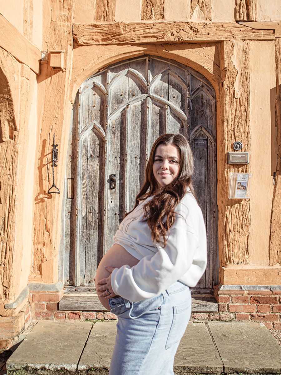 Mum‑to‑be in jeans and jumper cradling her bump in historic Lavenham market square, by all scene images photography. Essex, UK
