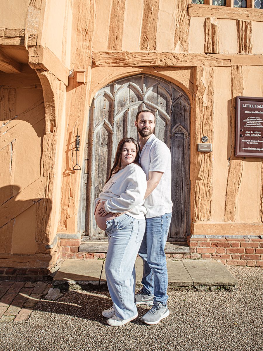 Mum‑to‑be in jeans and jumper cradling her bump in historic Lavenham market square, by all scene images photography. Essex, UK