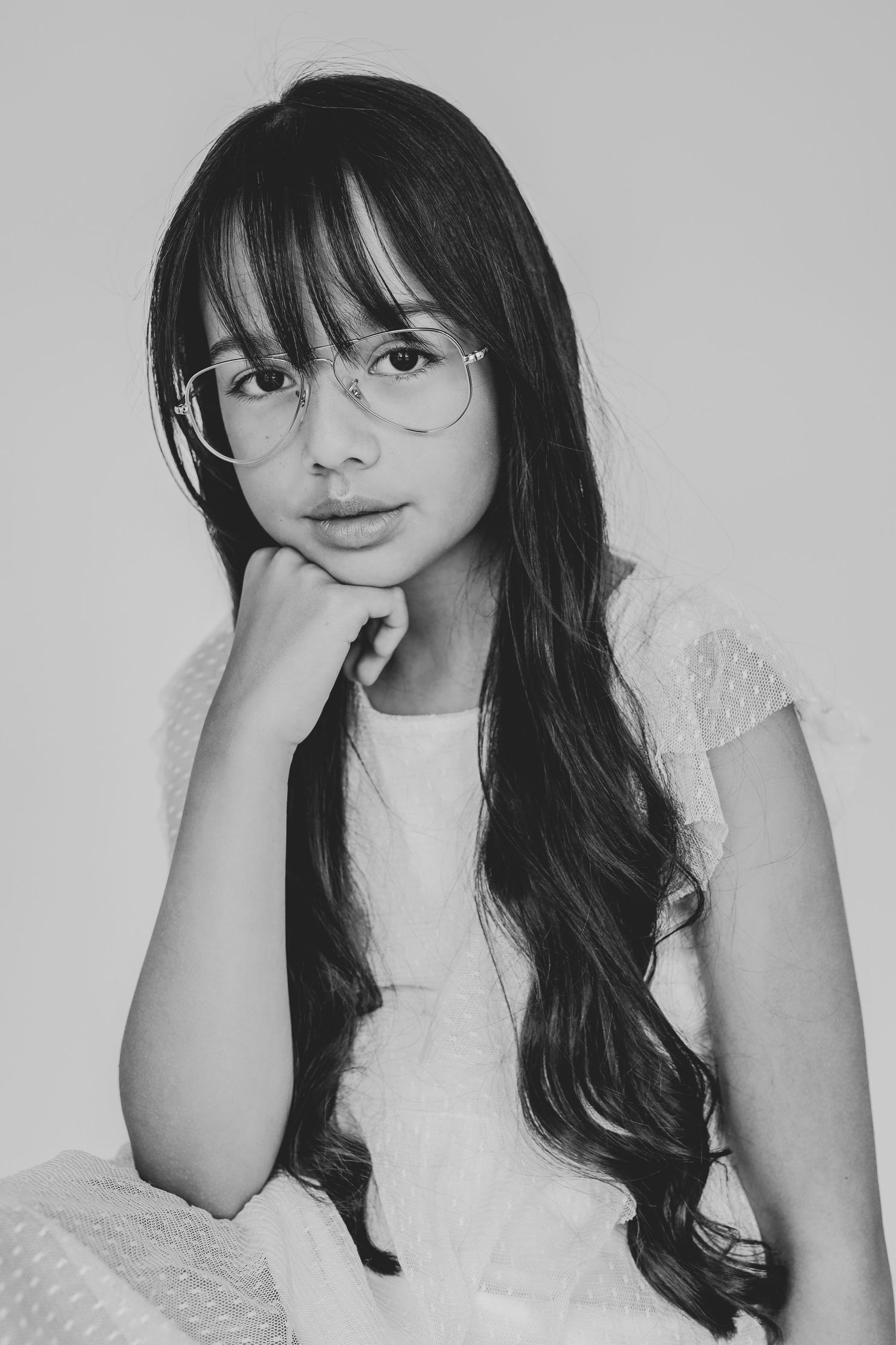 Black and white portrait of a young girl with glasses and long dark hair resting chin on hand.