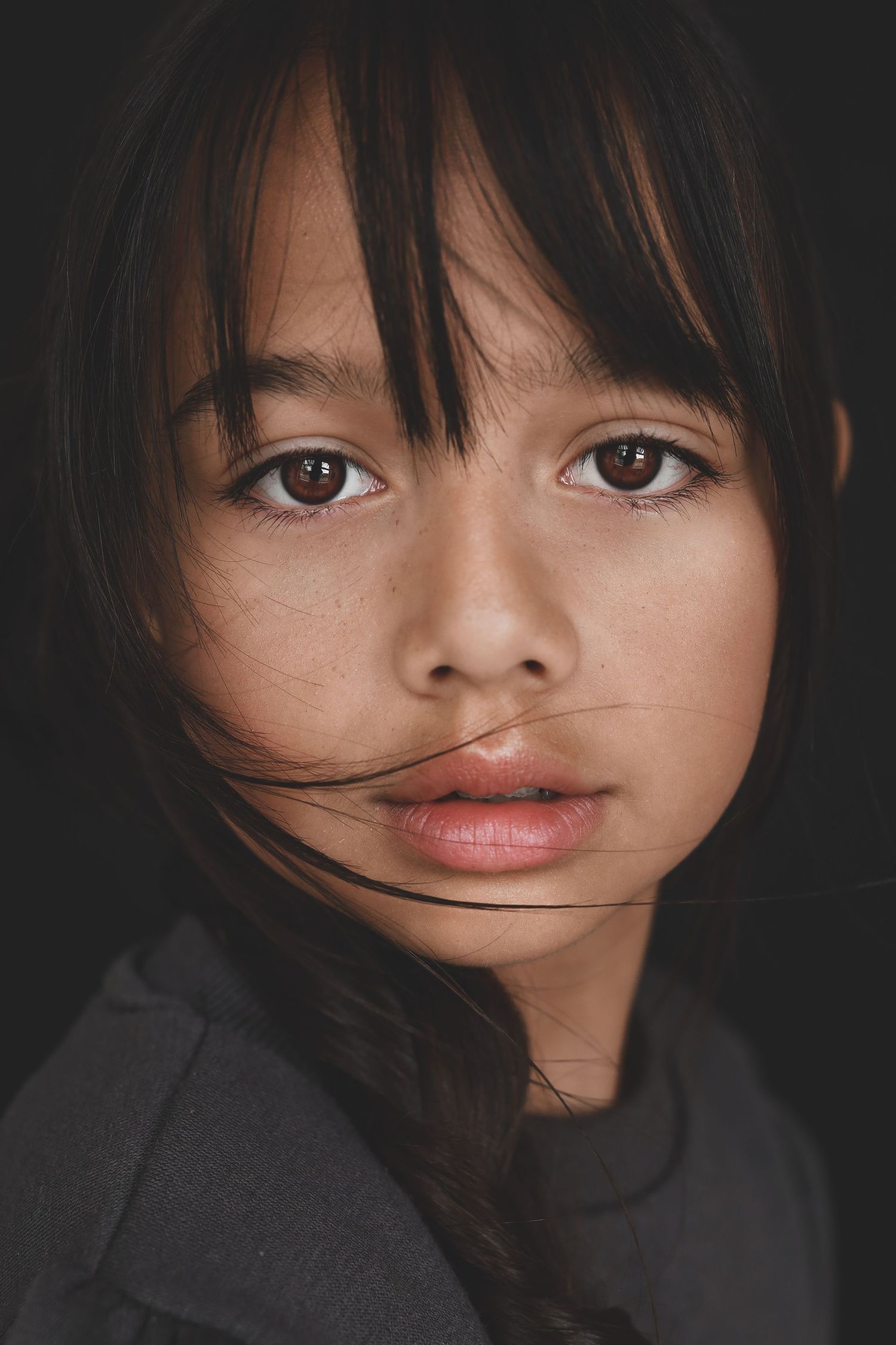 Close-up portrait of a young girl with dark bangs and brown eyes against a dark background.