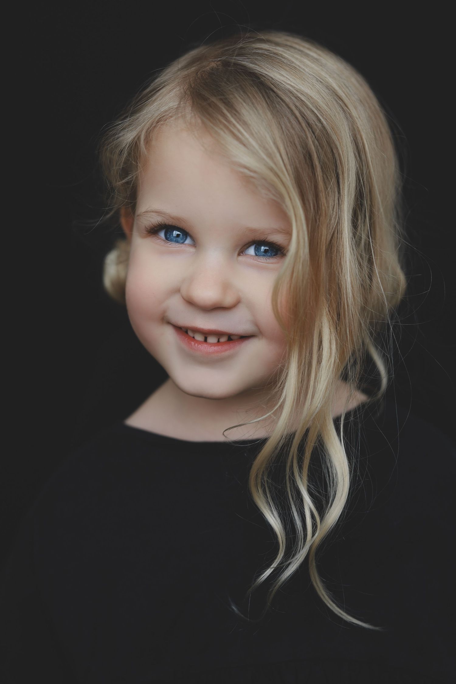 Smiling blonde little girl with blue eyes and long wavy hair against a dark background.
