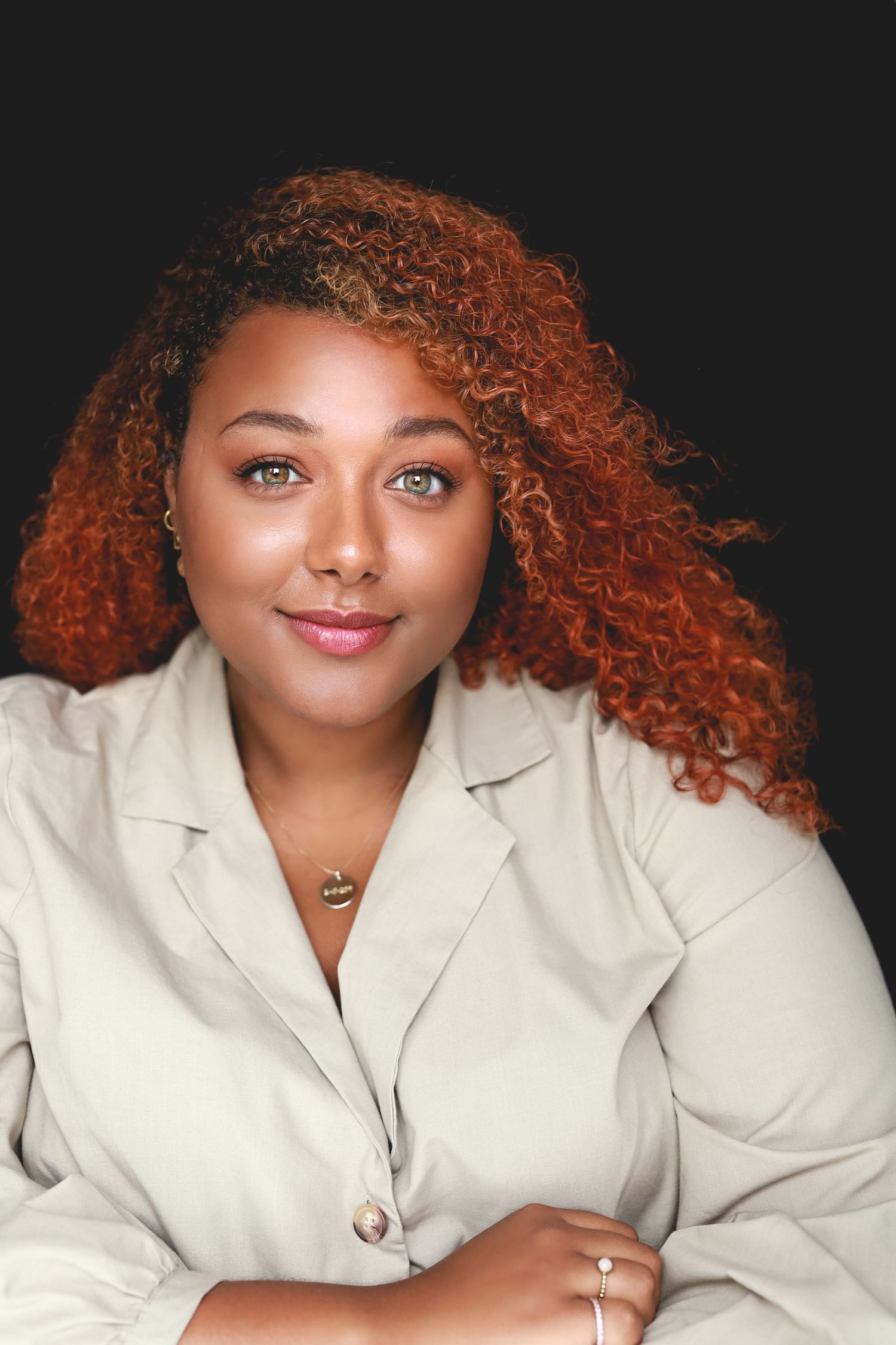 Woman with curly red hair and green eyes smiling confidently in a beige blazer.