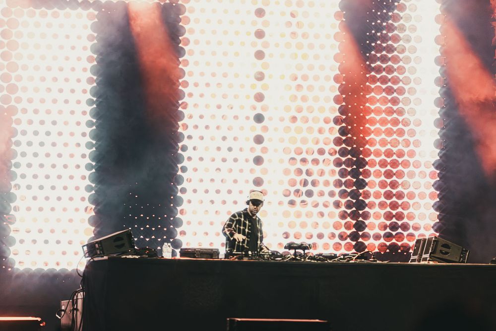 DJ performs on stage with dramatic red and white light display behind mixing console at music festival.