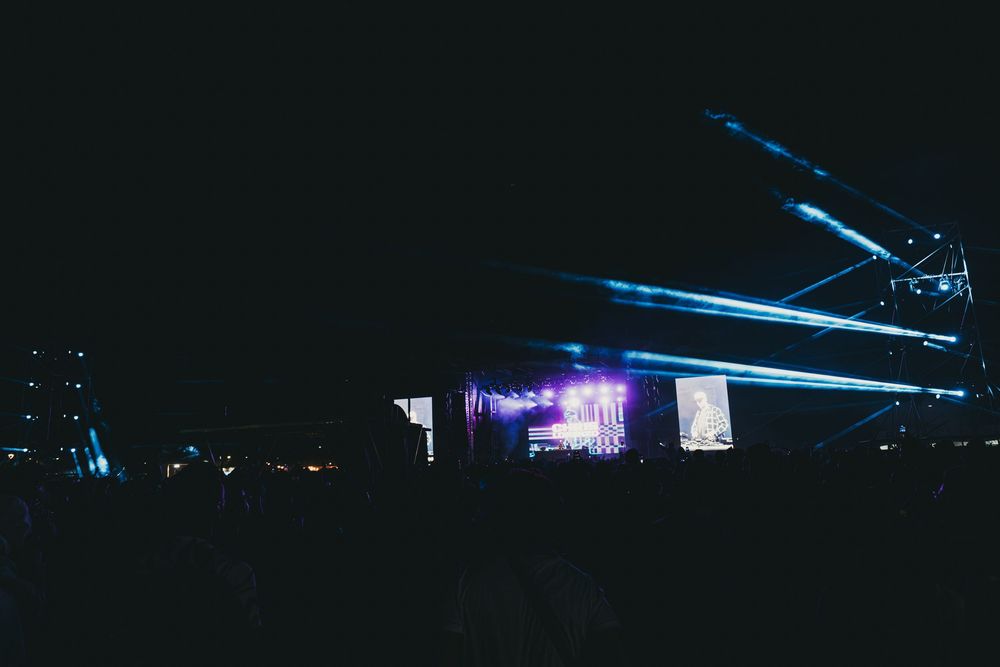 Blue laser beams illuminate a dark outdoor concert venue with a large video screen display glowing in the distance.