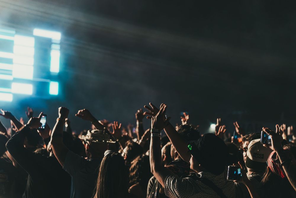 Concert crowd with raised hands silhouetted against bright stage lights during a live music performance.