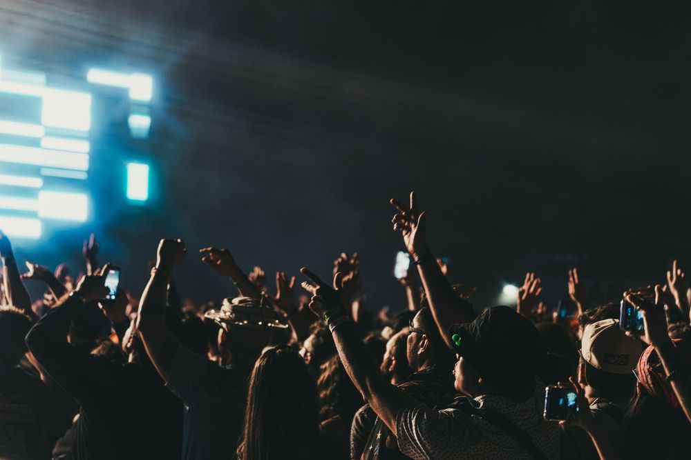 Concert crowd with raised hands silhouetted against bright stage lights during a live music performance.