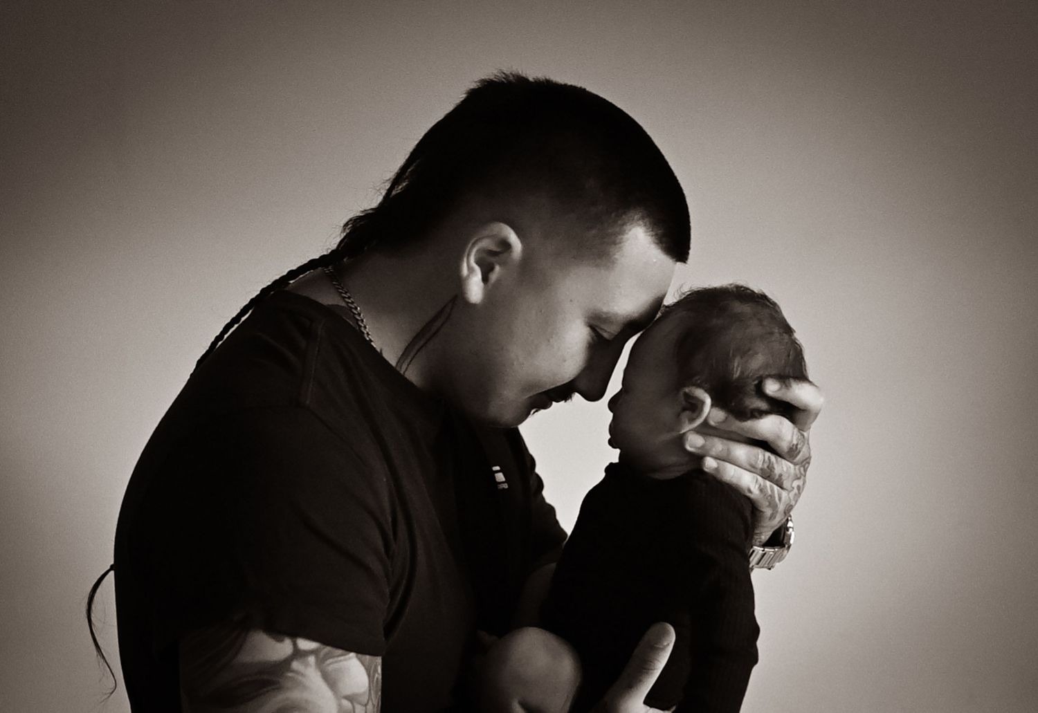 Sepia-toned photo of a tattooed father tenderly touching foreheads with his newborn baby.