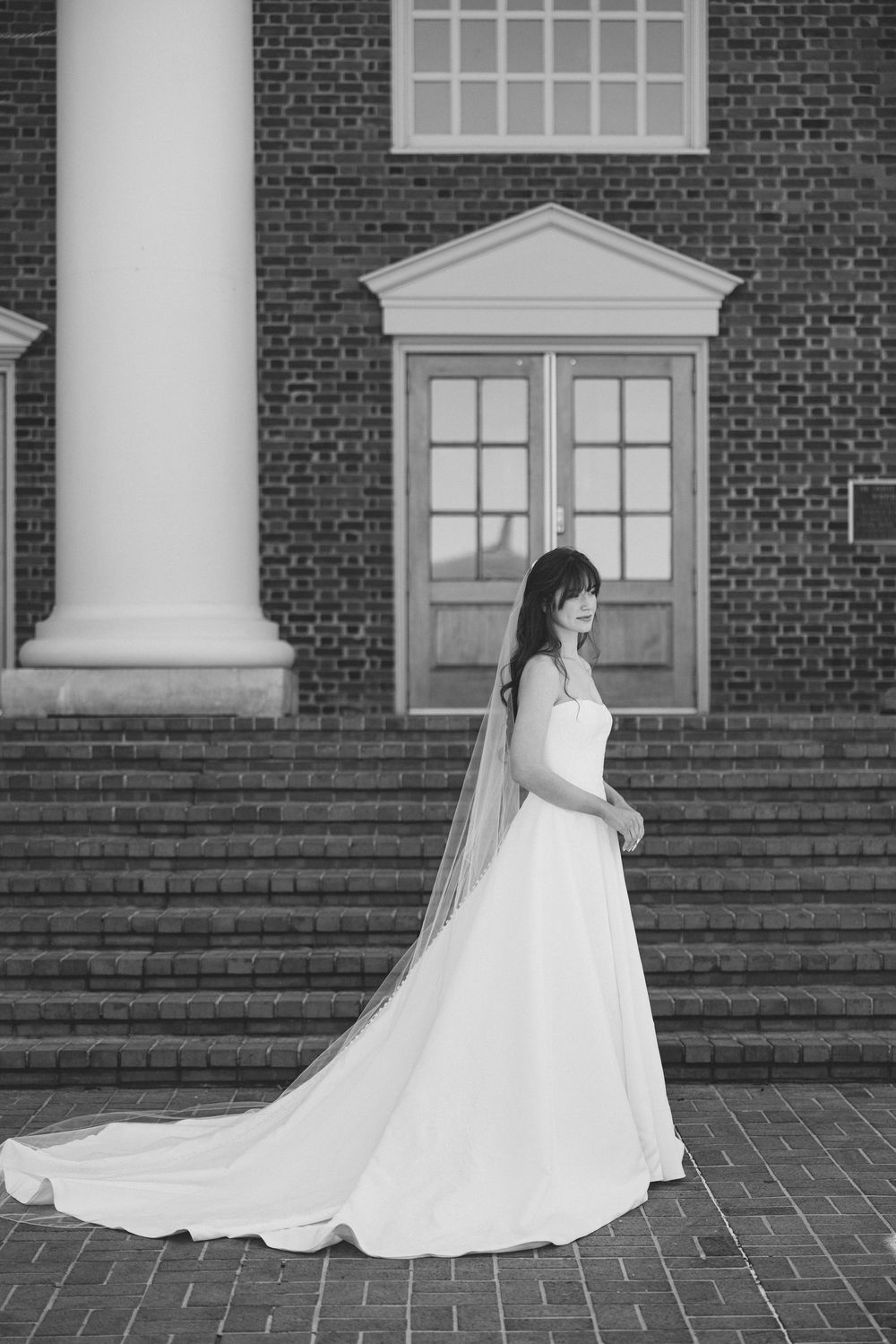 A bride in a flowing white wedding gown poses on brick steps in front of a classic colonial building entrance.