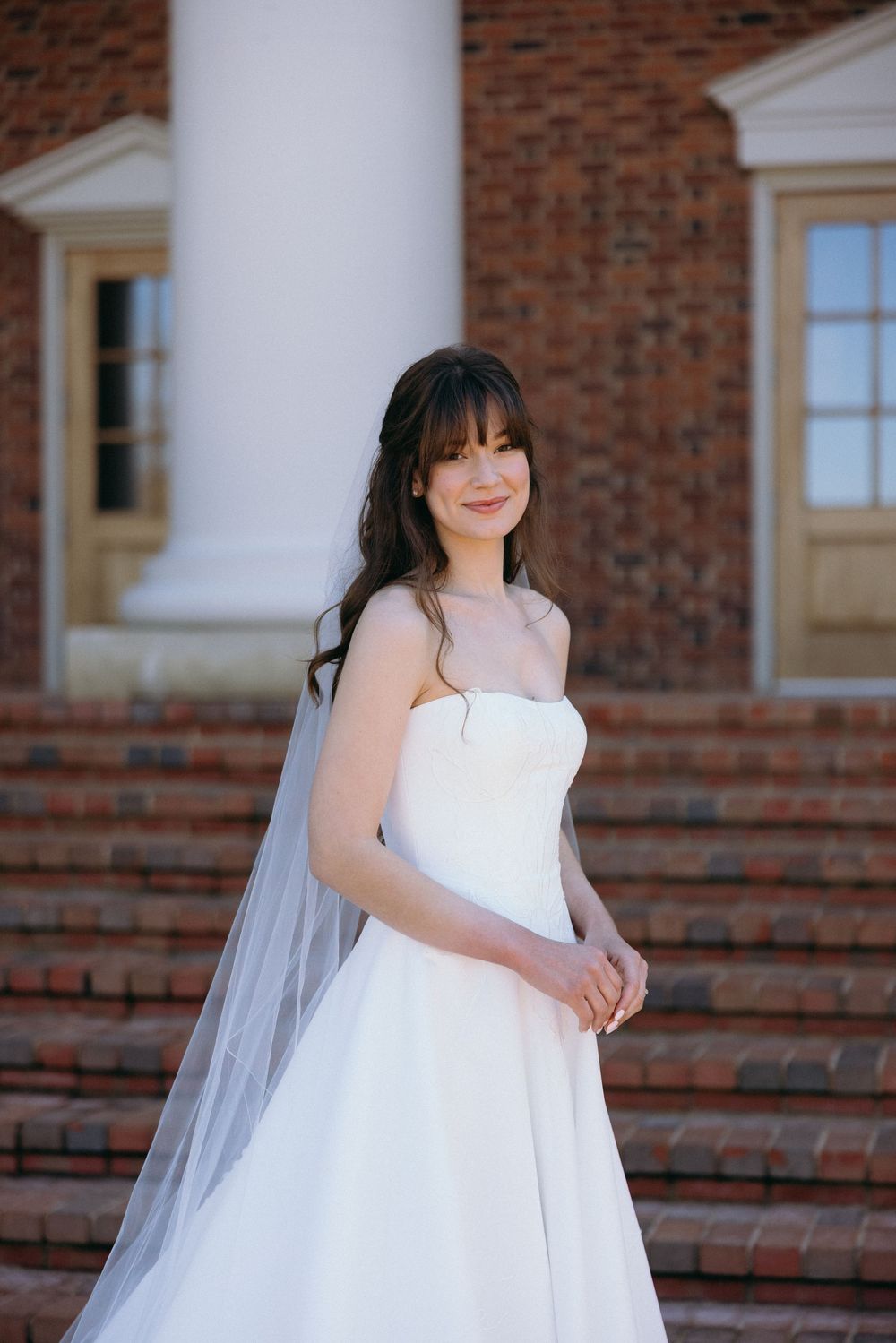 A bride in a white wedding dress poses in front of a brick building with white trim details.